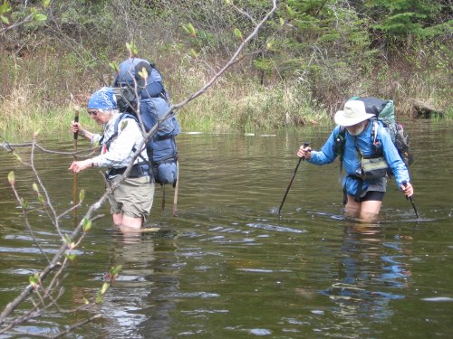 Two hikers wade through shallow water, carrying large backpacks and using trekking poles, surrounded by greenery.