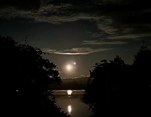A serene night scene with a full moon reflecting on a calm lake, framed by dark silhouettes of trees and clouds.