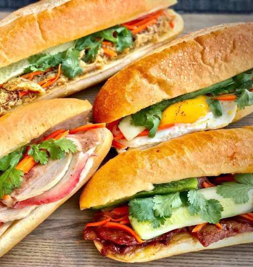 A variety of colorful sandwiches with fresh vegetables and herbs, arranged on a wooden surface.