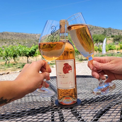 Three glasses of wine toast over a bottle of Oak Creek wine, set against a vineyard backdrop.