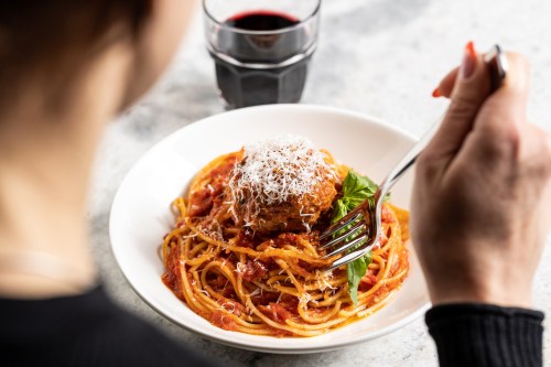 A person holds a fork, ready to eat spaghetti with meatball and cheese, accompanied by a glass of red wine.