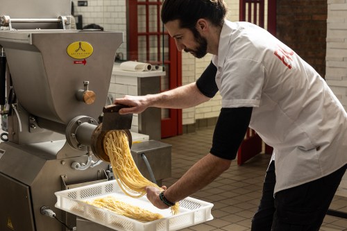 A chef in a white shirt operates a pasta machine, pouring freshly made spaghetti into a white basket.