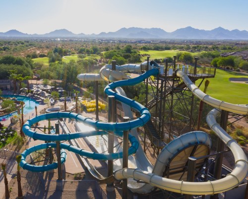 Aerial view of colorful water slides at a resort, surrounded by greenery and mountains in the background.
