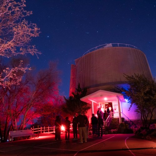 A group of people gathers outside a dome observatory under a starry night sky, illuminated by red lights.