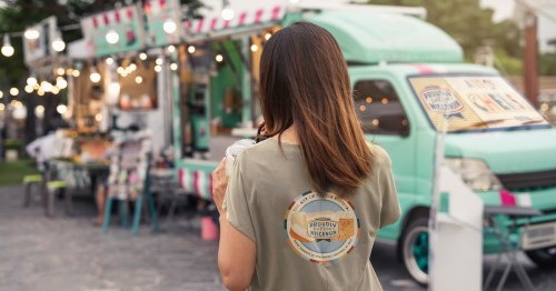 A woman stands with her back to the camera, holding a drink, in front of a colorful food truck at an outdoor market.