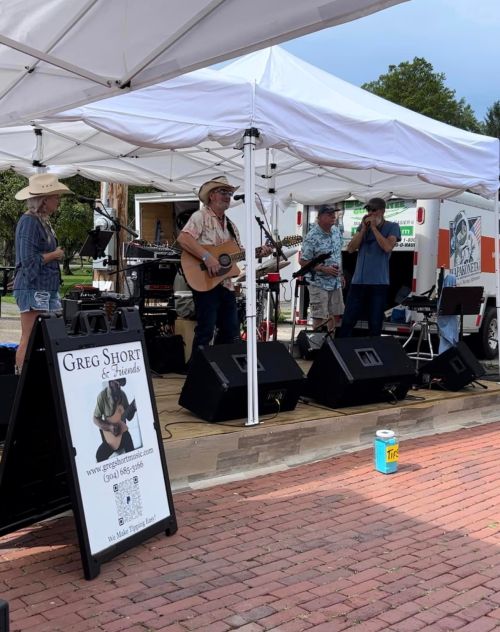A band performs under a tent at an outdoor event, with instruments and a sign for "Greg Short & Friends" visible.