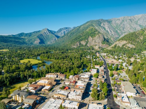 Aerial view of a small town surrounded by mountains and greenery, with a river and clear blue sky in the background.