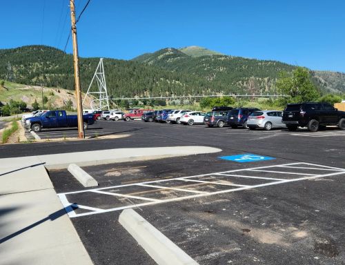 A parking lot with various cars, surrounded by green mountains and clear blue skies. A power line runs in the background.