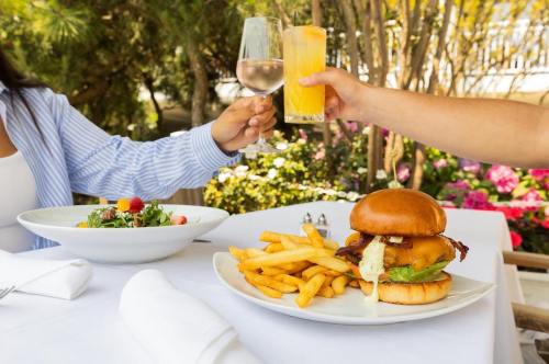 A table set with a burger, fries, a salad, and drinks, with two people toasting in a garden setting.