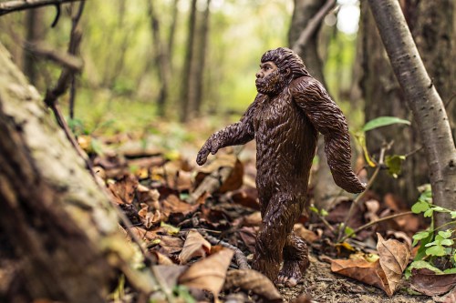 A brown Bigfoot figurine walking through a forest with fallen leaves and trees in the background.