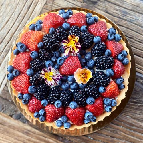 A colorful fruit tart topped with strawberries, blueberries, blackberries, and edible flowers on a wooden surface.