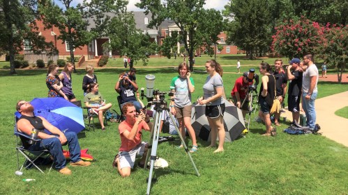 A group of people set up telescopes and equipment on a grassy area, some sitting under umbrellas, enjoying a sunny day.