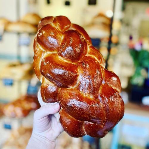 A hand holding a shiny, braided loaf of challah bread in a bakery setting.