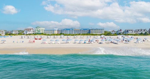 A sunny beach scene with umbrellas, lounge chairs, and a hotel in the background under a blue sky with fluffy clouds.