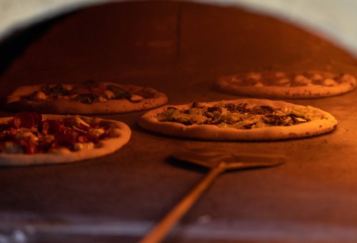 Four pizzas baking in a wood-fired oven, with a pizza peel resting nearby.