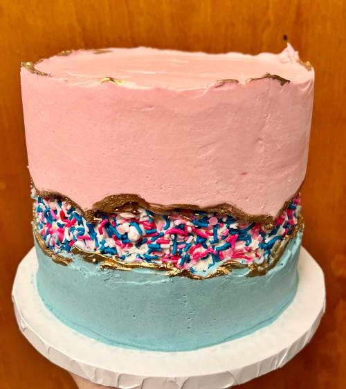 A three-layer cake with pink, blue, and white frosting, decorated with colorful sprinkles and a gold edge.