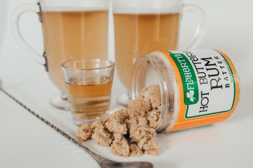 Two glasses of frothy drinks beside a shot glass and a spilled jar of Hot Buttered Rum batter.