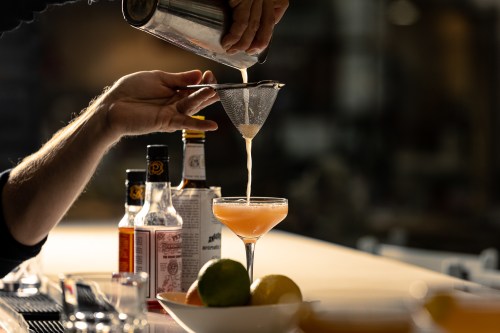 A bartender strains a cocktail into a glass, surrounded by bottles and fresh ingredients on a bar counter.