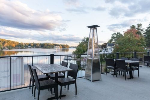 Outdoor dining area with tables and chairs overlooking a calm lake and trees, under a cloudy sky.