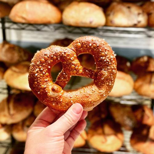 A hand holds a heart-shaped pretzel in front of a display of various baked goods.