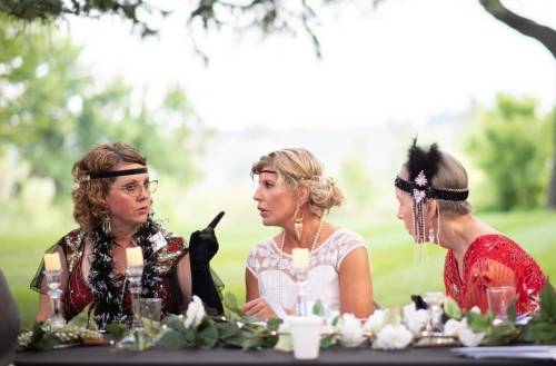 Three women in vintage attire engaged in a lively conversation at an outdoor table decorated with flowers and candles.