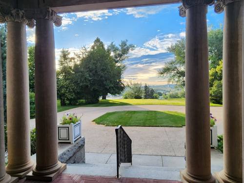 View from a porch framed by columns, showcasing a landscaped garden and distant hills under a blue sky.