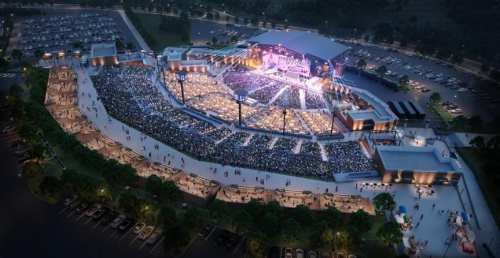 Aerial view of a large outdoor concert venue filled with a crowd, surrounded by parking and trees.