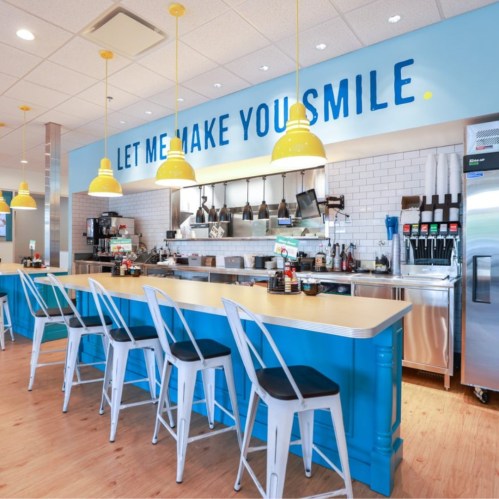 Bright café interior with blue counters, yellow pendant lights, and seating, featuring a cheerful slogan on the wall.