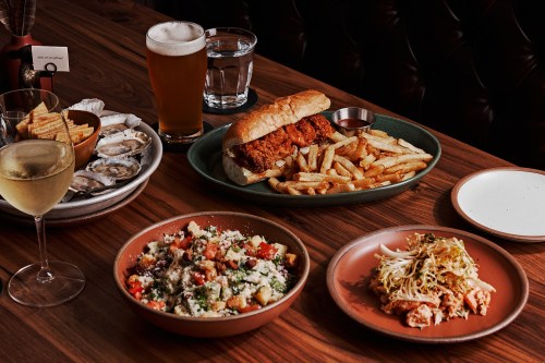 A table spread with fried chicken sandwich, fries, salads, oysters, and drinks, set in a cozy dining atmosphere.