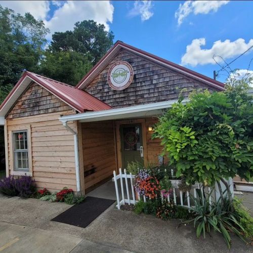 A charming wooden building with a red metal roof, surrounded by colorful flowers and greenery, under a blue sky.