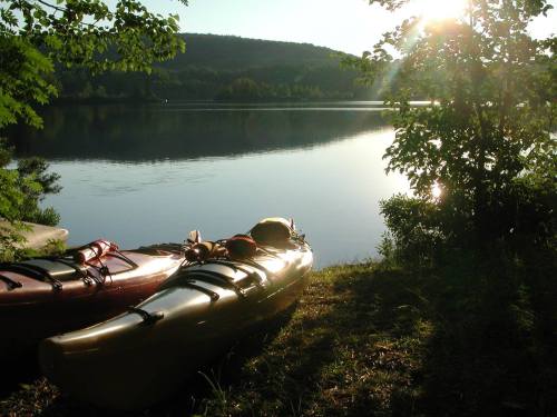 Two kayaks on the shore of a calm lake, surrounded by trees, with sunlight reflecting on the water.