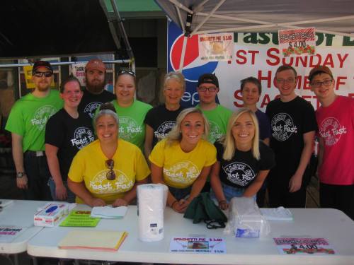 A group of people in colorful t-shirts smiling at a food festival booth, promoting a community event.