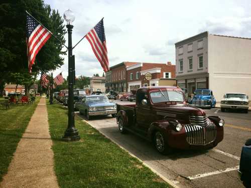 A vintage red truck parked on a street lined with classic cars and American flags, in a small town setting.