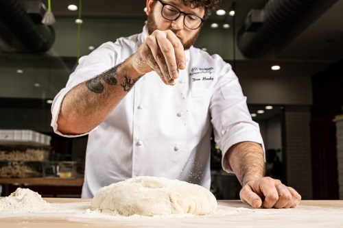A chef in a white coat sprinkles flour over a dough on a wooden surface, focused on his baking process.