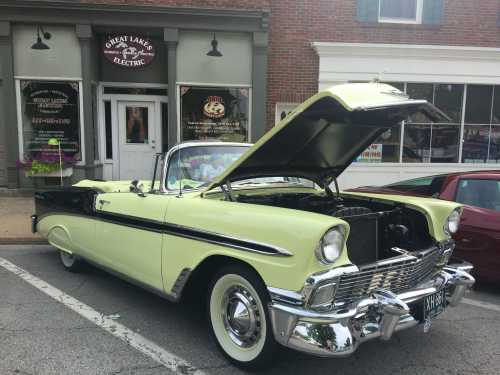 A vintage yellow convertible car with the hood open parked in front of a brick building with a sign for a bakery.