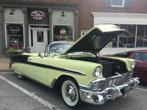 A vintage yellow convertible car with the hood open parked in front of a brick building with a sign for a bakery.