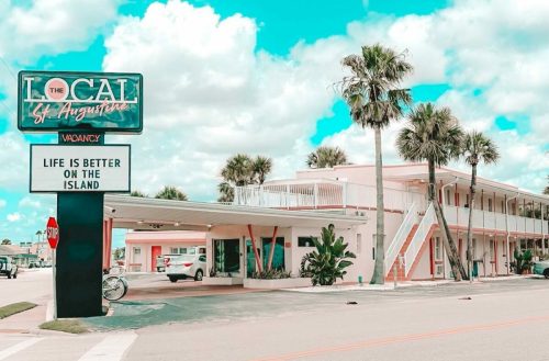 A pink hotel with palm trees and a sign reading "Life is better on the island" under a bright blue sky.