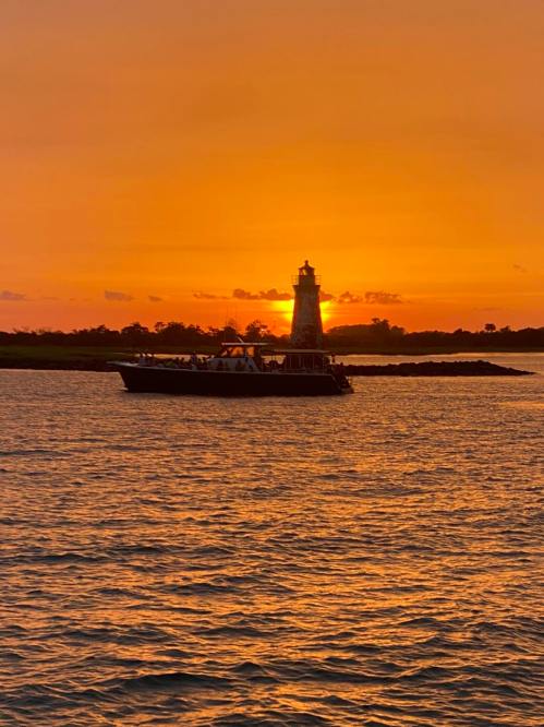 A silhouette of a lighthouse against a vibrant orange sunset, with a boat in the foreground on calm waters.