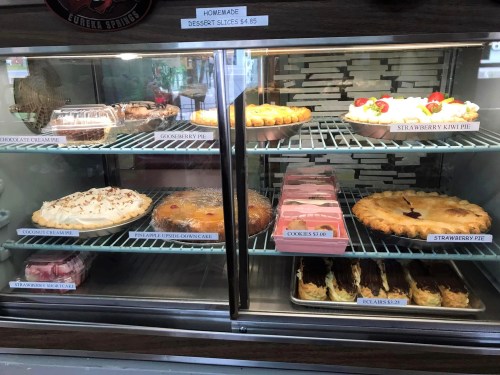 Display case filled with various desserts, including pies, cakes, and eclairs, with labels and prices visible.