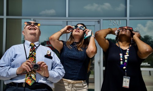 Three people wearing eclipse glasses look up, smiling, in front of a building on a sunny day.