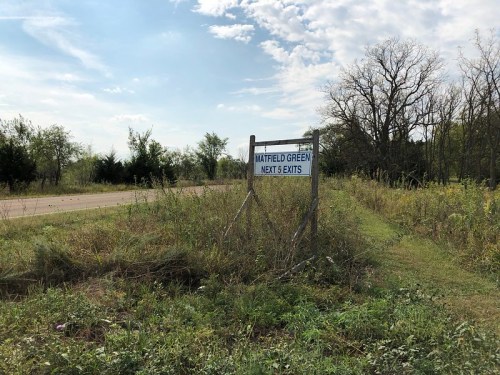 Sign for "Hatfield Green" with directions for the next 1 mile exits, surrounded by overgrown grass and trees.