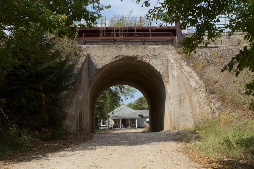 A concrete archway leads to a gravel path, with a house visible in the background and trees lining the sides.