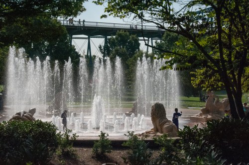 A fountain with water jets in a park, surrounded by greenery and a bridge in the background. People are enjoying the area.