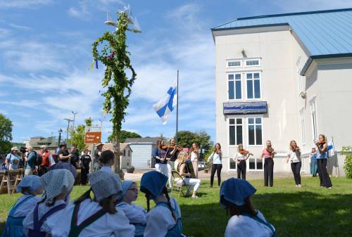 A group performs with violins near a Finnish flag, while children in traditional attire watch in a sunny outdoor setting.