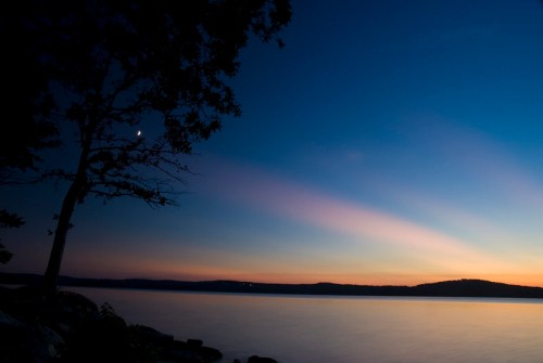 A serene lakeside scene at dusk, featuring a tree silhouette and a crescent moon against a colorful sky.