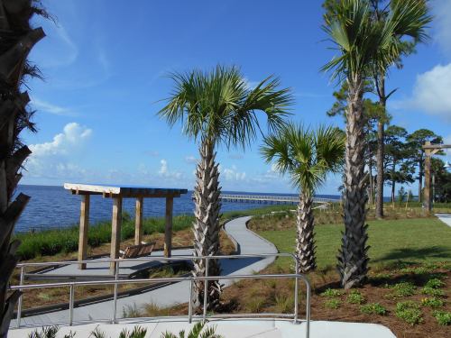 A scenic view of a waterfront park featuring palm trees, a wooden gazebo, and a pier under a blue sky.