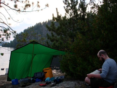 A person sits by a lake near a green tent, surrounded by camping gear and trees, with mountains in the background.