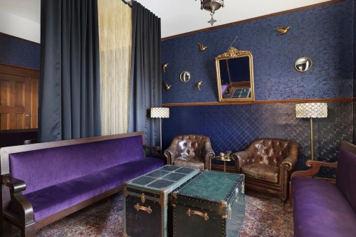 A cozy lounge with purple sofas, leather chairs, a vintage mirror, and decorative lamps against a dark blue wall.