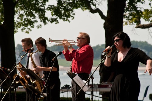 A lively band performs outdoors, featuring a singer, trumpet player, and saxophonist, with a scenic backdrop.