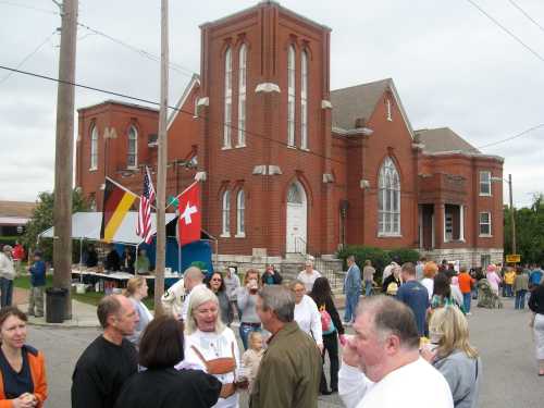 A crowd gathers outside a red brick church during a community event, with flags and tents in the background.